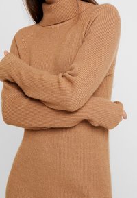 Pull à manches longues et col montant, en maille côtelée. Fabriqué en tissu doux d'une couleur beige chaud avec une silhouette ajustée.