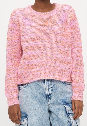 Person trägt einen strukturierten rosa-orangefarbenen Strickpullover kombiniert mit hellblauen, acid-washed Cargo-Jeans und steht vor einem schlichten Hintergrund.