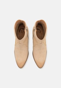 Botas de tobillo beige de ante con un diseño festoneado, punta afilada, cremalleras laterales y costuras en contraste a lo largo de la suela.