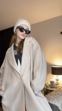 Világos bézs, túlméretezett kabát sállal, sötét pulóverre viselve. Fekete napszemüveggel és krémszínű kötött sapkával párosítva.