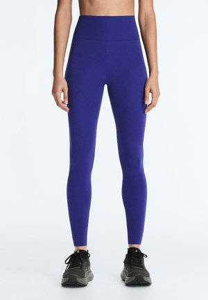 Kobieta ubrana w jasnoniebieskie legginsy z wysokim stanem i czarne buty sportowe, stojąca prosto na jednolitym białym tle.