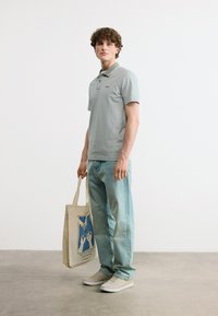 Jeune homme portant un polo gris et un jean bleu clair, tenant un sac fourre-tout beige avec un motif bleu, debout sur un sol gris.