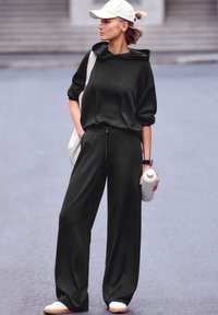 Tumši zaļš, brīvi pieguļošs džemperis ar kapuci un atbilstošas platas bikses, apavi ar baltām kedām un baltā ūdens pudele rokās.