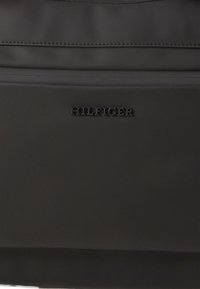 Crna najlonska torba s glatkom teksturom, slim dizajnom i sjajnim "HILFIGER" logom sprijeda. Vidljivo rubno šivanje duž šavova.