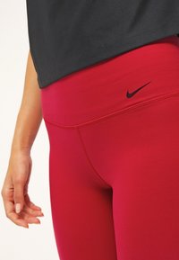 Leggings rossi realizzati in materiale elasticizzato, dotati di una vita alta e un piccolo logo Nike nero vicino alla parte superiore. Texture liscia con un design aderente.