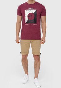 T-shirt in cotone borgogna con stampa grafica in bianco e nero, abbinato a pantaloni corti beige arrotolati. Il modello indossa sneakers bianche.