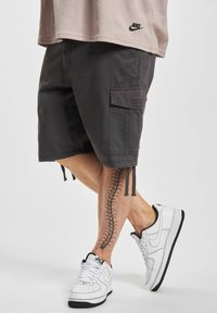 Schwarze knielange Cargo-Shorts mit Seitentaschen, Kordelzügen und glatter Textur. Getragen mit weißen Nike-Sneakern und einem beigen Oberteil.