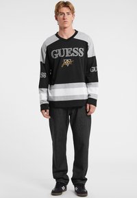 Svart och grå långärmad tröja med "GUESS" text och logotyp. Har randigt mönster och V-ringning. Bärs med mörka jeans och sneakers.