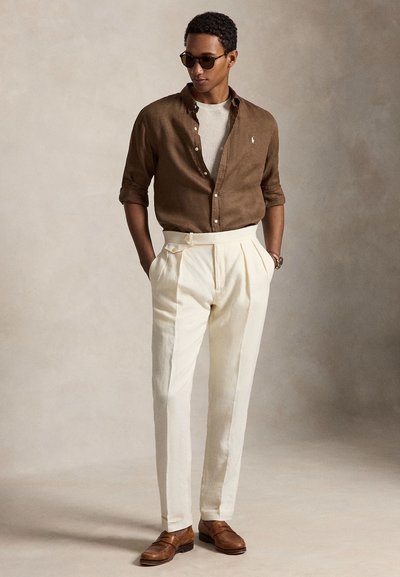 Bărbat purtând o cămașă băiețoasă maro peste un top alb, pantaloni plisati crem, mocasini maro și ochelari de soare, stând cu mâinile în buzunare.