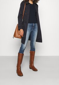 Marinblå trenchcoat, kabelstickad marinblå tröja, blå jeans, bruna läderstövlar och en brun axelväska. Stövlarna har sidodragkedjor och remmar.