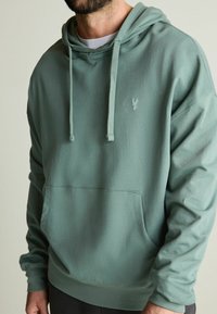Mikina s kapucí z měkké, tlumené zelené látky. Má přední kapsu, šňůrky a malé vyšívané logo na hrudi.