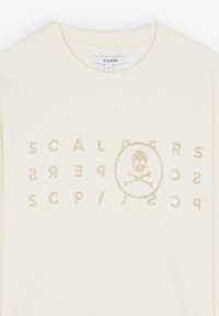 Sweatshirt couleur crème avec un col rond, présentant un logo en métal texturé doré et un design de skull sur le devant. Coupe classique.