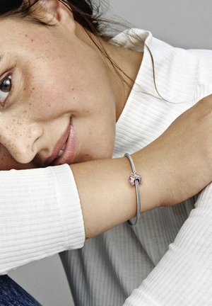 Silbernes Charm-Armband mit einem pinken Akzent, strukturiertem Design und flexibler Kette, getragen an einem Handgelenk mit einem weißen gerippten Oberteil.