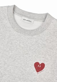 Pelēks sporta krekls ar ribotu apļa apkakli. Izceļas ar sarkanu sirds grafiku ar smaidu un tekstu "OH APRIL" apakšā. Mīksta tekstūra.