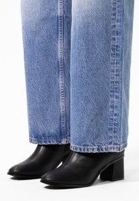 Bottines noires en cuir à talon avec un bout carré, talon carré épais, associées à un jean en denim bleu clair avec une texture décolorée et une coupe droite.
