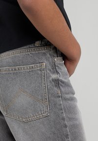 Pantalones vaqueros de mezclilla gris con costuras visibles, detalles en los bolsillos traseros y un ligero desvanecimiento. La persona lleva una camisa negra.
