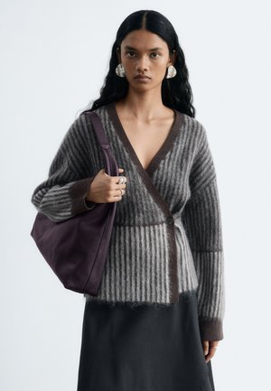 Donna con lunghi capelli neri indossa grandi orecchini d’argento, un cardigan grigio a righe peloso, una gonna nera e porta una borsa a tracolla in pelle viola scuro.