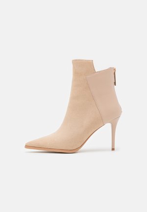 Bottine beige à talon haut avec bout pointu, avant en suède, arrière en cuir lisse, et fermeture éclair au talon.