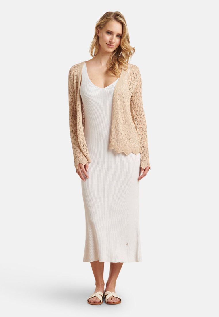 Beige gebreid vest over een witte geribde jurk met een aangesloten lijfje en uitlopend zoom. De sandalen zijn crèmekleurig met een gestructureerde band.