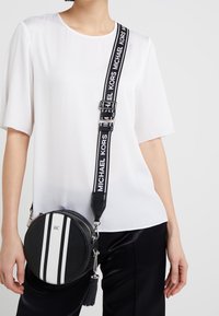 Borsa a tracolla rotonda realizzata in pelle nera texture, con strisce bianche, accessori in argento e un dettaglio a frange sulla cerniera.