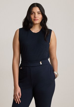 Femme aux longs cheveux foncés portant un haut noir sans manches et un pantalon noir taille haute avec des boutons, debout devant un fond uni.