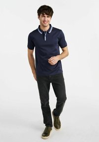 Granatowa koszulka polo z kontrastującymi detalami kołnierzyka, noszona z ciemnymi dżinsami i zielonymi trampkami. Materiał jest gładki, a krój casualowy i dopasowany.
