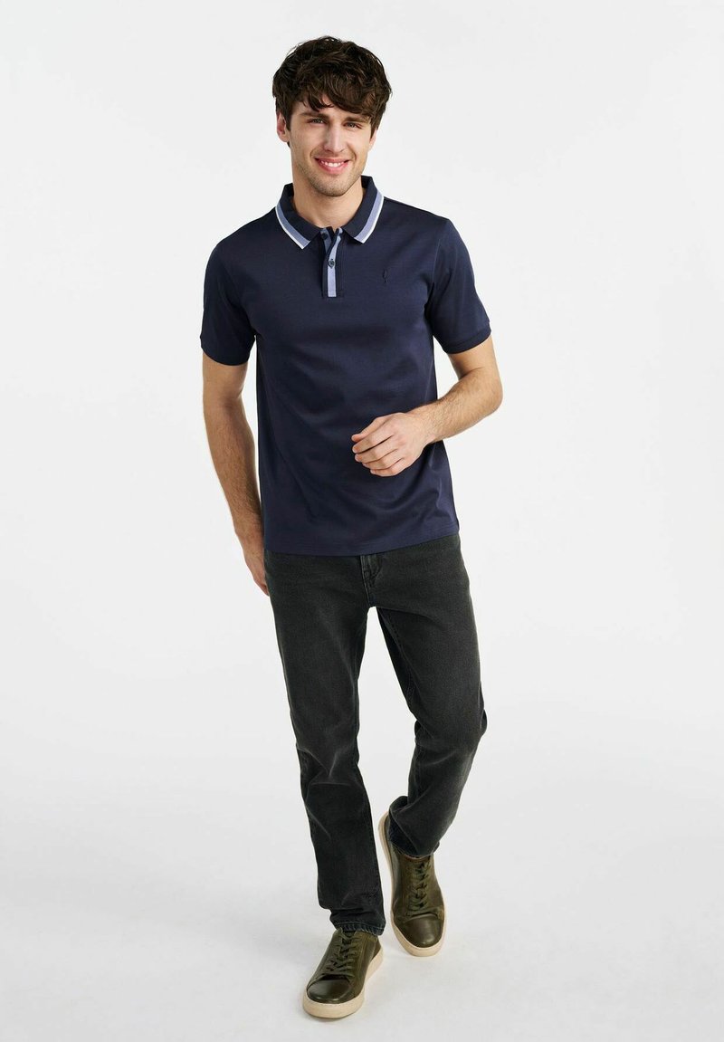 Granatowa koszulka polo z kontrastującymi detalami kołnierzyka, noszona z ciemnymi dżinsami i zielonymi trampkami. Materiał jest gładki, a krój casualowy i dopasowany.
