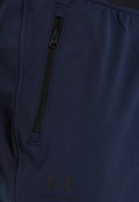 Närbild av marinblått tyg med en svart vertikal dragkedjeficka och Under Armour-logotypen under fickan.