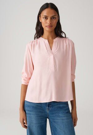 Hellrosa Bluse mit einem geschlitzten Ausschnitt, langärmlig und lässig hochgekrempelt, aus einem glatten Stoff, kombiniert mit blauen Jeans aus Denim.