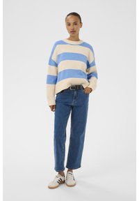 Femeie în picioare, purtând un pulover rayat crem și albastru, blugi cu centură neagră și adidași albi cu dungi negre.