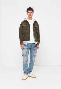 Young man standing against white background wearing olive jacket over gray hoodie, white t-shirt, light blue cargo jeans, and white sneakers.