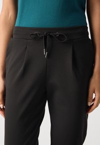 Schwarze Hose mit Kordelzugbund, seitlichen Falten und silbernem Zubehör. Der Stoff wirkt glatt und hat einen entspannten Schnitt.