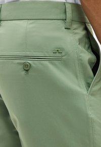 Nærbillede af lysegrønne bukser, der viser baglommes knap, sidelomme, bæltestrop og lille logodetalje på stoffet.