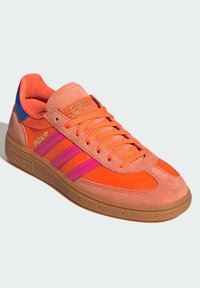 Orange suede sneaker with pink stripes, gum sole, and blue accent on the heel. Features lace-up design and padded tongue for comfort.