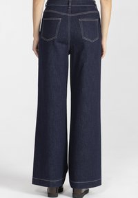 Osoba nosząca ciemnoniebieskie dżinsy o szerokich nogawkach i czarne buty na platformie, pokazana od tyłu na tle białego tła.