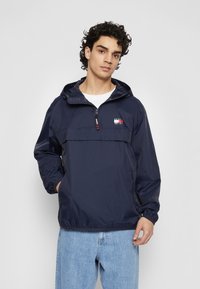Navy blue hooded windbreaker featuring a half-zip front, elastic cuffs, and a mid-chest logo patch. Made of lightweight, shiny nylon material.