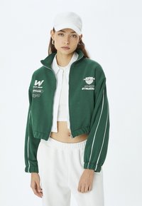 Chaqueta corta verde con cremallera y acentos de rayas blancas, que presenta logotipos bordados y un cuello alto. Combinada con pantalones blancos de cintura elástica.