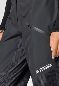 Calças exteriores pretas com tecido padronizado na lateral, com um bolso com fecho éclair e um logo "TERREX" visível em branco.