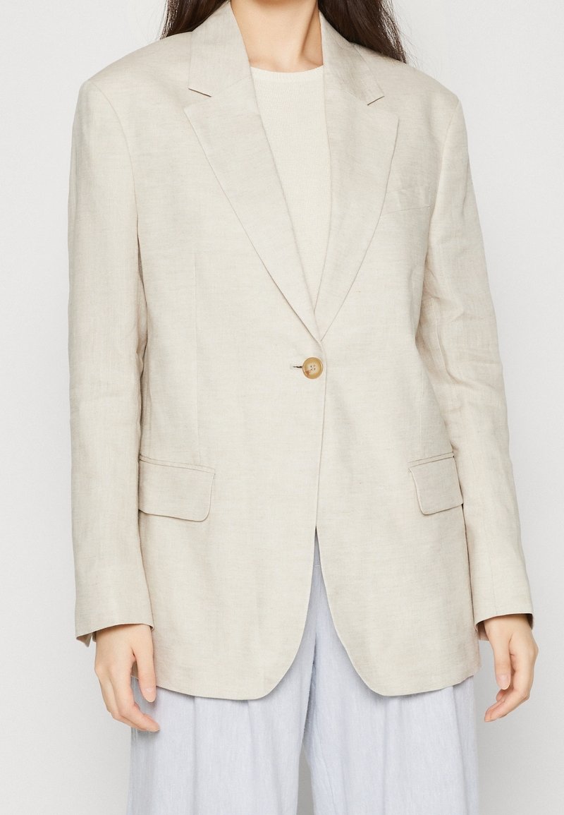 Béžový lněný blejzr s jedním knoflíkem, klopovými límci a dvěma předními kapsami. Materiál má texturovaný a lehký vzhled.