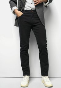 Mann, der schwarze Hosen, weiße Sneakers, ein helles Hemd und ein graues Sakko trägt, steht mit einer Hand in der Tasche vor einem weißen Hintergrund.