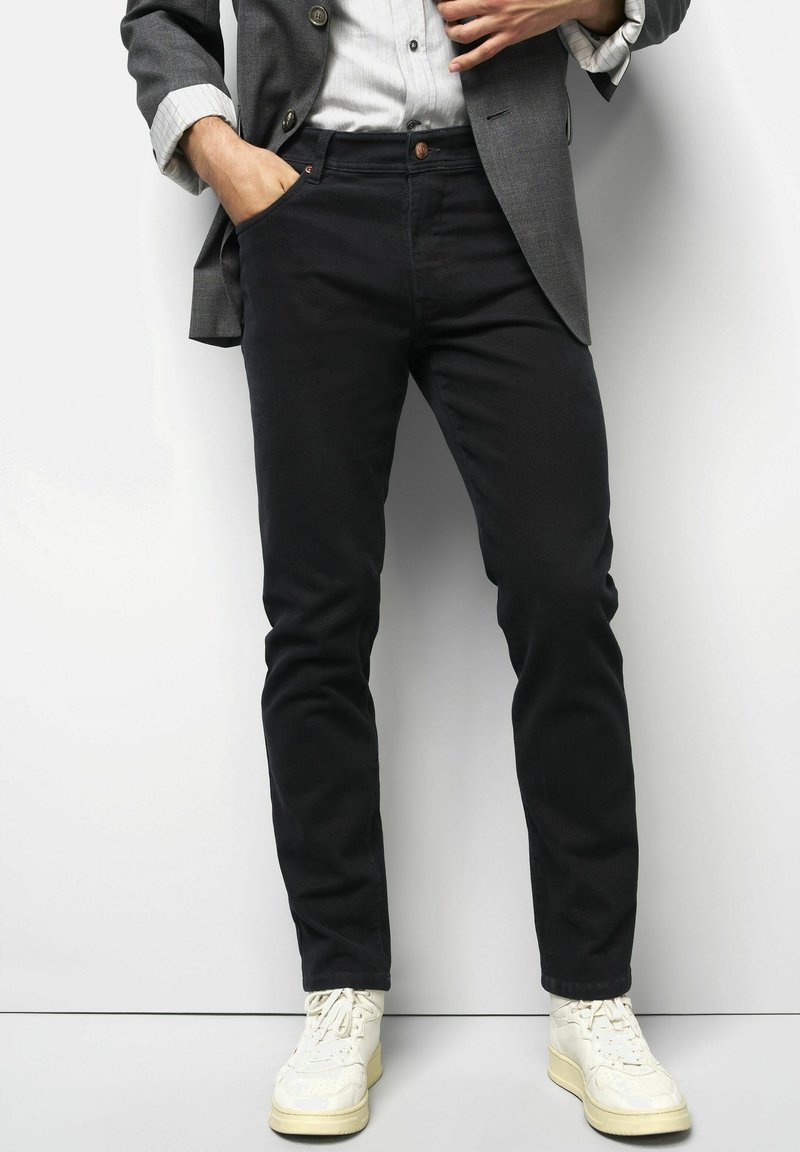 Mann, der schwarze Hosen, weiße Sneakers, ein helles Hemd und ein graues Sakko trägt, steht mit einer Hand in der Tasche vor einem weißen Hintergrund.