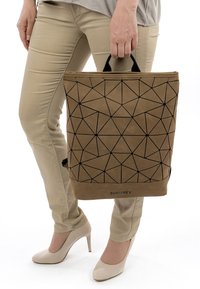 Persona con pantalones beige y tacones sosteniendo un bolso Suri Frey marrón con patrón geométrico por su asa negra.