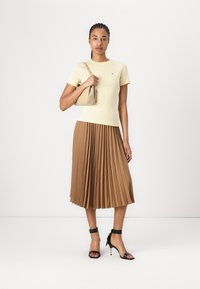Light yellow fitted t-shirt with short sleeves, paired with a pleated brown midi skirt. Model holds a beige handbag, wearing black strappy heels.