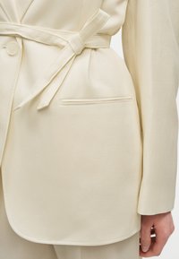 Gros plan sur un blazer crème avec ceinture en tissu nouée, bouton unique, poche passepoilée et manches longues, porté avec un pantalon assorti.