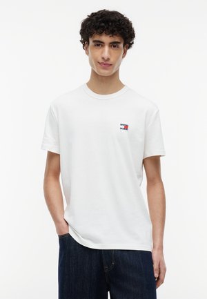 Junger Mann mit lockigem Haar, der ein weißes T-Shirt mit kleinem Logo auf der Brust und dunkelblaue Jeans trägt, stehend mit einer Hand in der Tasche.