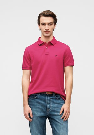 Tânăr care poartă un tricou polo roz aprins cu mâneci scurte și blugi albaștri, stând pe un fundal simplu și deschis la culoare.