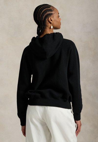 Vrouw met cornrows, gekleed in een witte broek en een zwart capuchontrui, met de rug naar de kijker tegen een neutrale achtergrond.