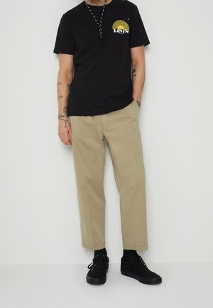 T-shirt nera con grafica gialla, pantaloni larghi beige e sneakers nere. Presenta tatuaggi sulle braccia e si trova in piedi davanti a uno sfondo neutro.