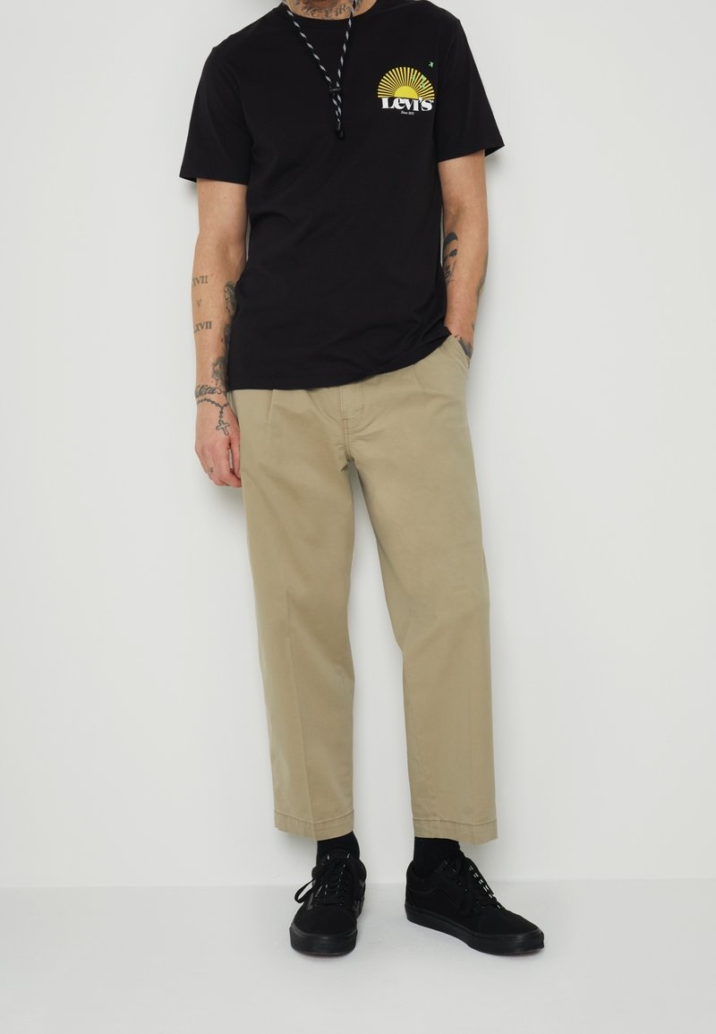 Camiseta negra con gráfico amarillo, pantalones anchos color beige y zapatillas negras. Muestra tatuajes en los brazos, de pie contra un fondo neutro.