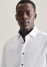 Junger schwarzer Mann trägt ein weißes Hemd mit schwarzen Knöpfen, schaut nach vorne vor einem schlichten hellen Hintergrund.